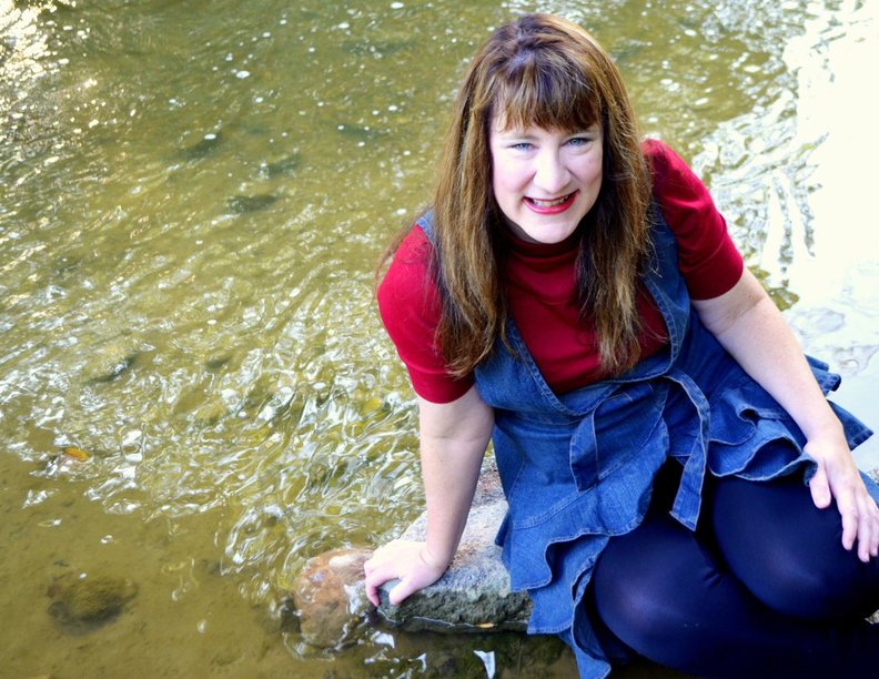 woman in a red top and blue skirt sitting on a stone in a river